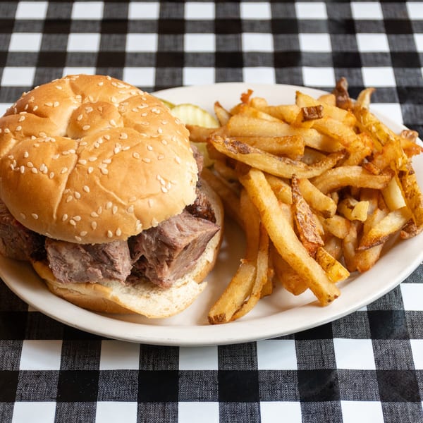 Brisket sandwich with crispy fries and pickles on a white plate, on a checkered tablecloth.