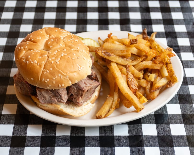 Brisket sandwich on a sesame bun with golden fries and pickles on a checkered tablecloth.