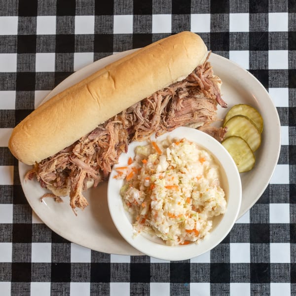 Pulled pork sandwich with coleslaw and pickles on a white plate over a black and white checkered tablecloth.
