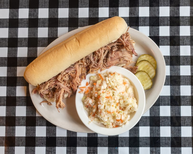 Pulled pork sandwich with coleslaw and pickles on a white plate over a black and white checkered tablecloth.