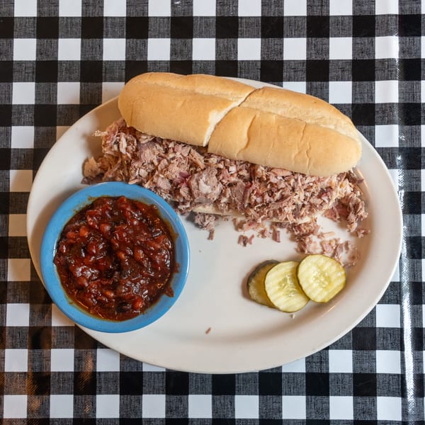 Pulled pork sandwich on a white plate with baked beans and pickles on a checkered tablecloth.