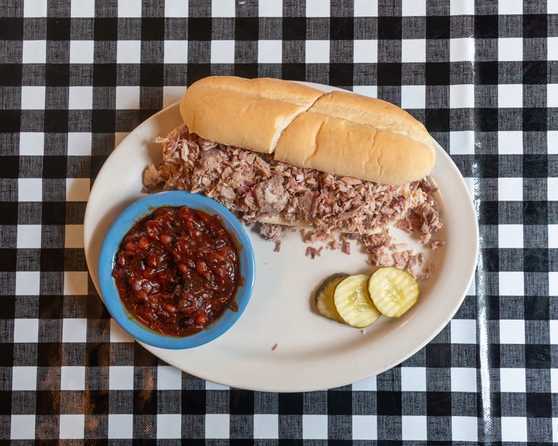 Pulled pork sandwich on a white plate with baked beans and pickles on a checkered tablecloth.