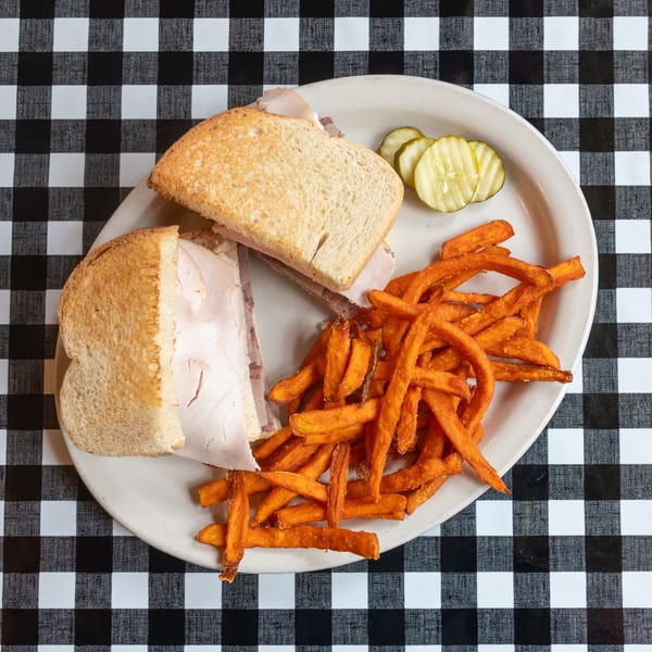 Deli meat sandwich with sweet potato fries and pickles on a white plate over a checkered tablecloth.