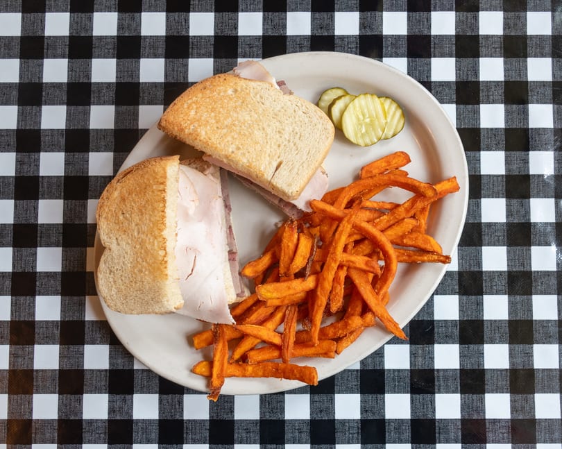 Turkey sandwich with sweet potato fries and pickles on a white plate over a checkered tablecloth.