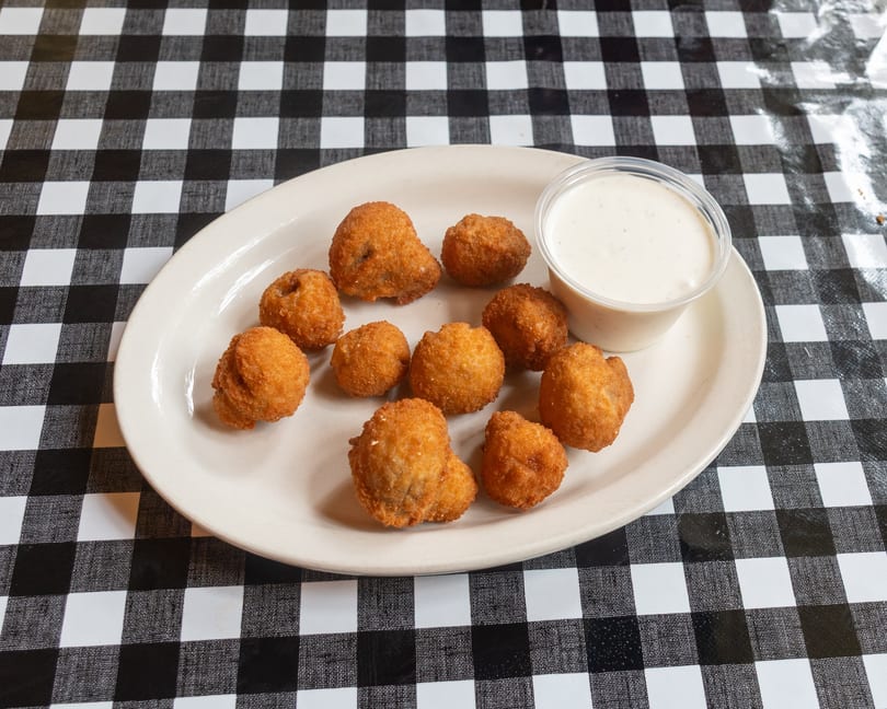 Golden-brown fried mushrooms and a cup of dipping sauce on a white plate over a checkered tablecloth.