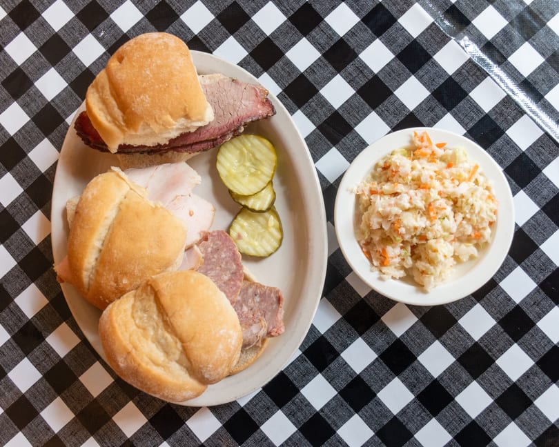 Deli sandwiches with various meats, pickles, and coleslaw on a black and white checkered tablecloth.