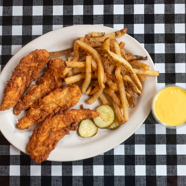 A plate of crispy chicken tenders, golden fries, pickles, and yellow sauce on a checkered table.