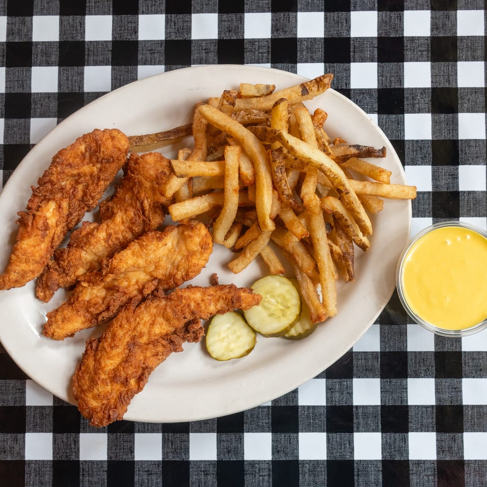 A plate of crispy chicken tenders, golden fries, pickles, and yellow sauce on a checkered table.