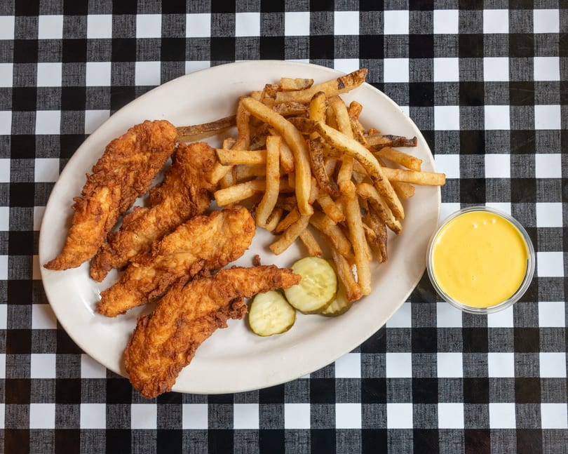 Crispy chicken tenders, french fries, pickles, and yellow dipping sauce on a checkered tablecloth.