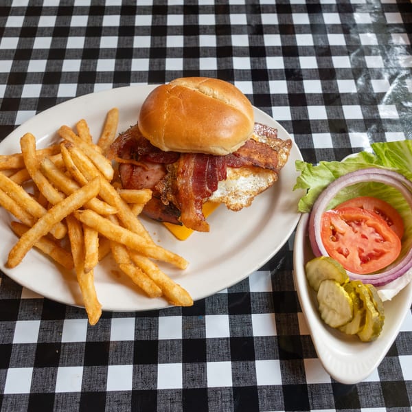 Bacon and egg burger with fries, lettuce, tomato, onion, and pickles on a checkered tablecloth.