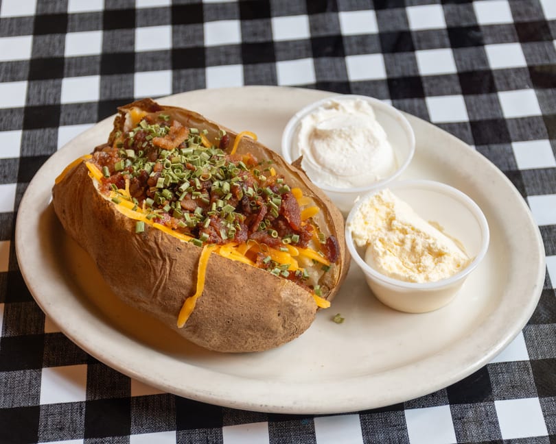 Loaded baked potato with cheese, bacon, chives, and sides of sour cream and butter on a checkered tablecloth.