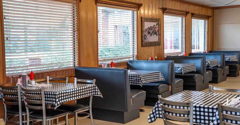Diner interior with black booths, checkered tablecloths, and wood-paneled walls with window blinds.