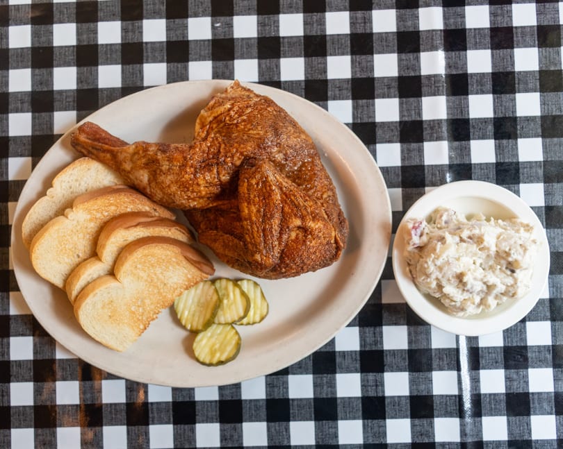 Roasted chicken leg and thigh with sliced bread, pickles, and potato salad on a checkered tablecloth.