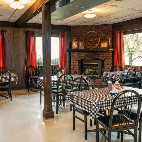 Empty dining room with checkered tablecloths, black chairs, red curtains, and a brick fireplace.