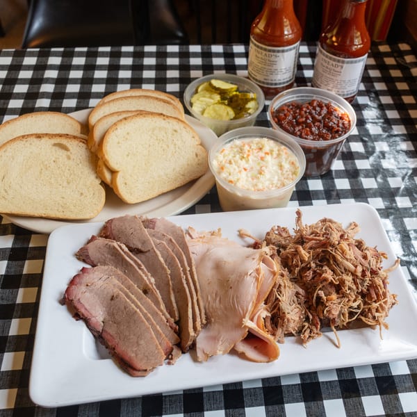 Platter of sliced brisket, turkey, and pulled pork with sides, bread, and BBQ sauce on a checkered tablecloth.