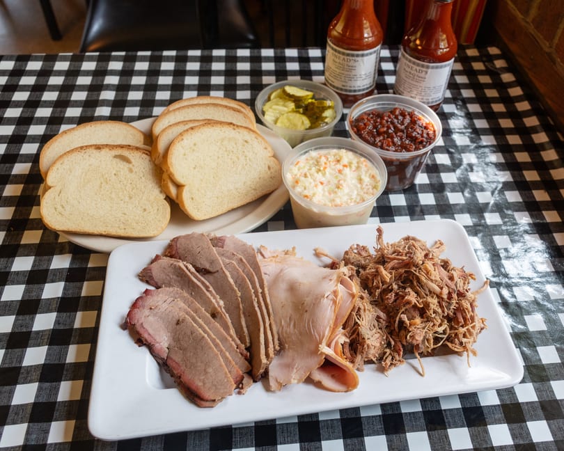Barbecue platter with sliced brisket, turkey, pulled pork, bread, coleslaw, beans, pickles, and sauce bottles.