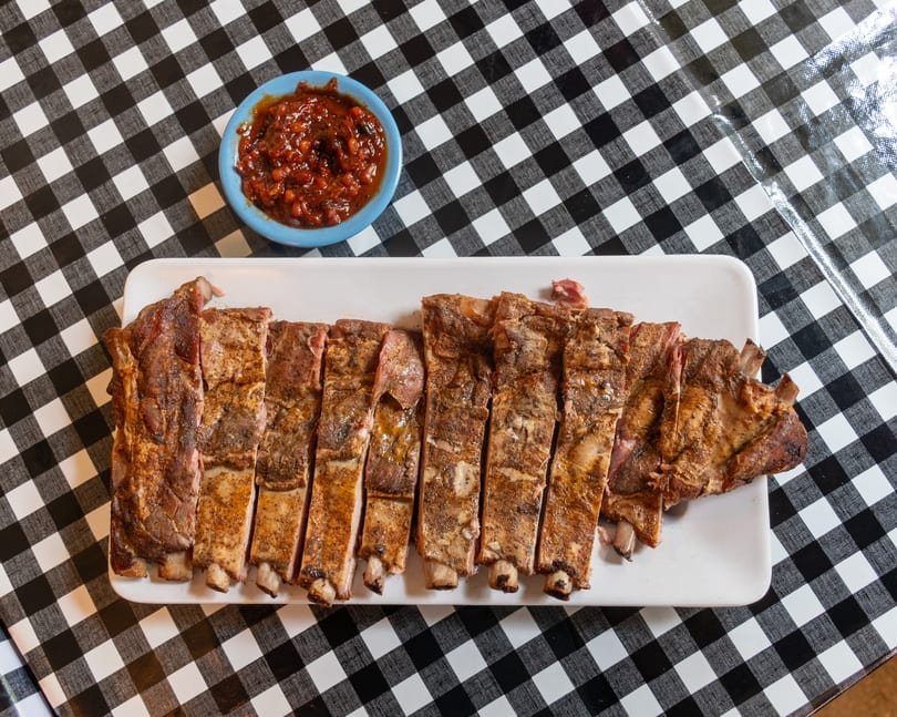 Sliced BBQ ribs on a white platter with a side of baked beans on a black and white checkered tablecloth.