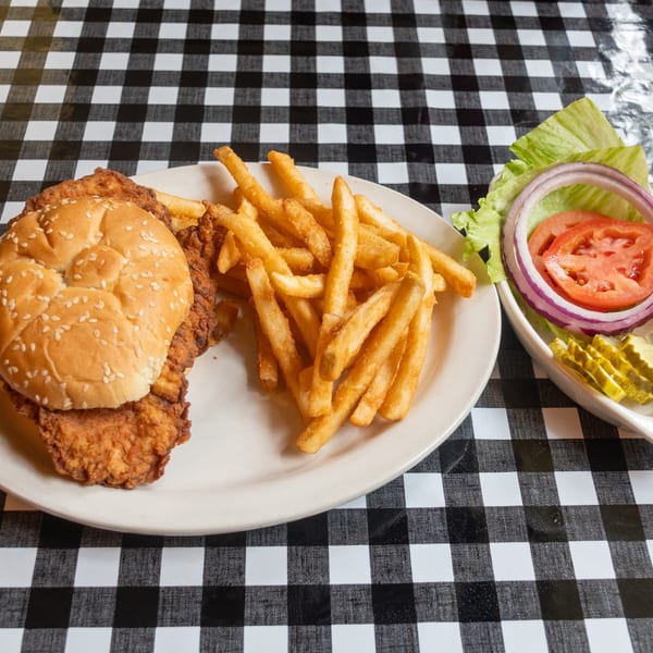 Fried chicken sandwich with fries and a side of lettuce, tomato, onion, and pickles on a checkered tablecloth.