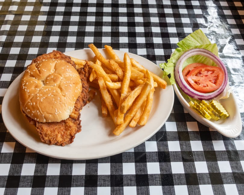 Fried chicken sandwich with fries and a side of lettuce, tomato, onion, and pickles on a checkered tablecloth.