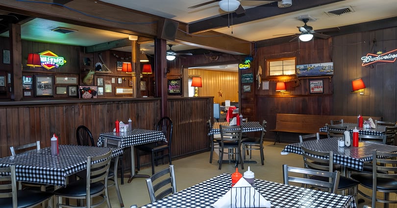 Cozy restaurant interior with checkered tablecloths, wood paneling, and neon signs on the walls.