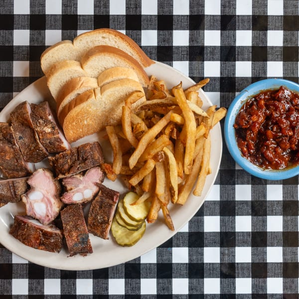 Plate of BBQ ribs, fries, bread, pickles, and baked beans on a checkered tablecloth.