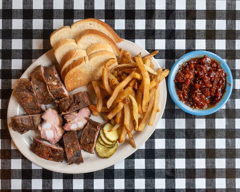 Plate of ribs, fries, bread, and pickles with a side of baked beans on a checkered tablecloth.